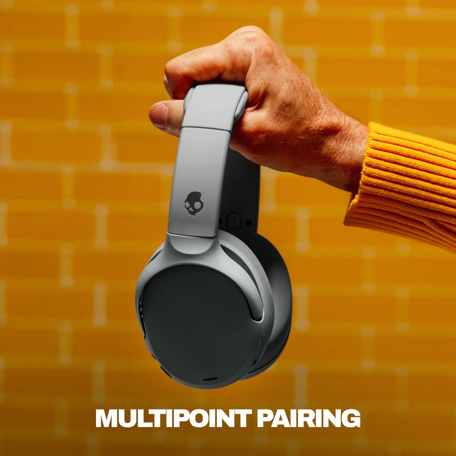 Hand holding a pair of Crusher 720 headphones with 'MultiPoint Pairing' text on a yellow brick wall background | Cement