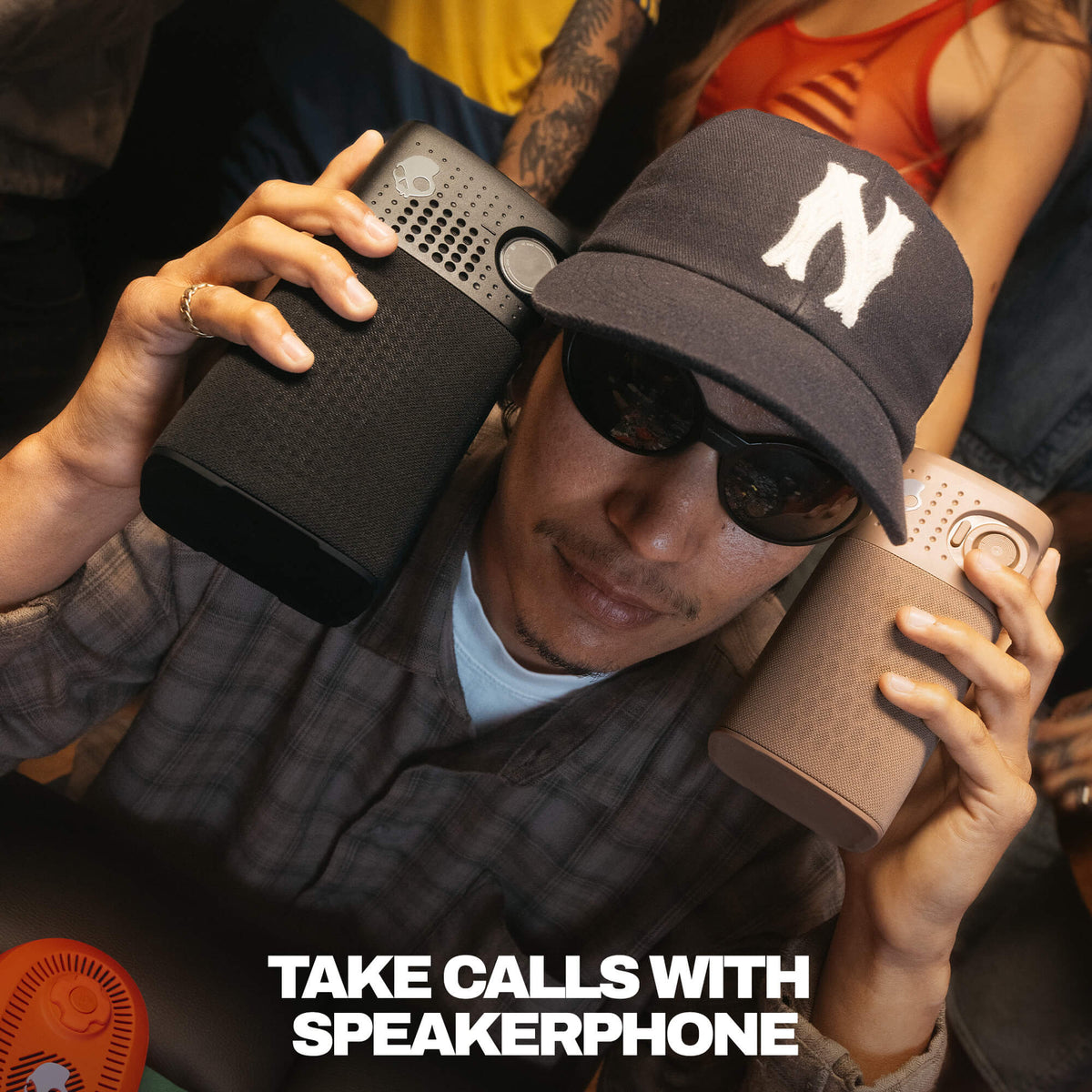 A man wearing a black baseball cap with a white 'N' logo, sunglasses, and a plaid shirt holds two session 900 speakerphones, one black and one taupe, to his ears. The text 'TAKE CALLS WITH SPEAKERPHONE' is displayed at the bottom. | Primer