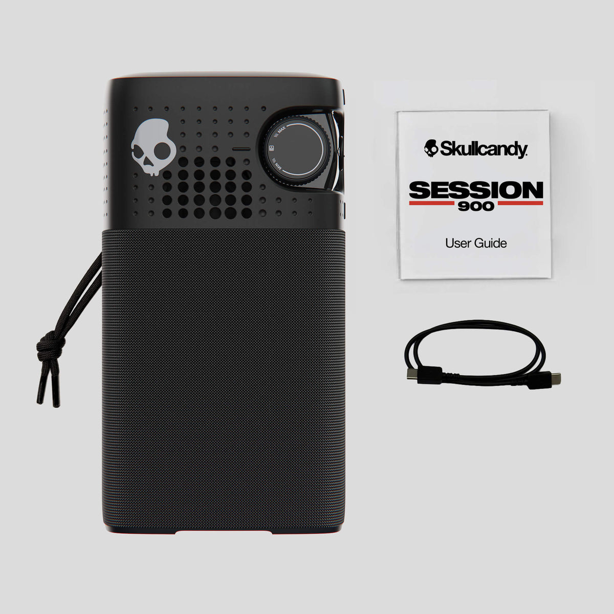 A black Skullcandy Session 900 portable speaker shown with its user guide booklet, charging cable, and carrying strap on a light gray background. | Primer