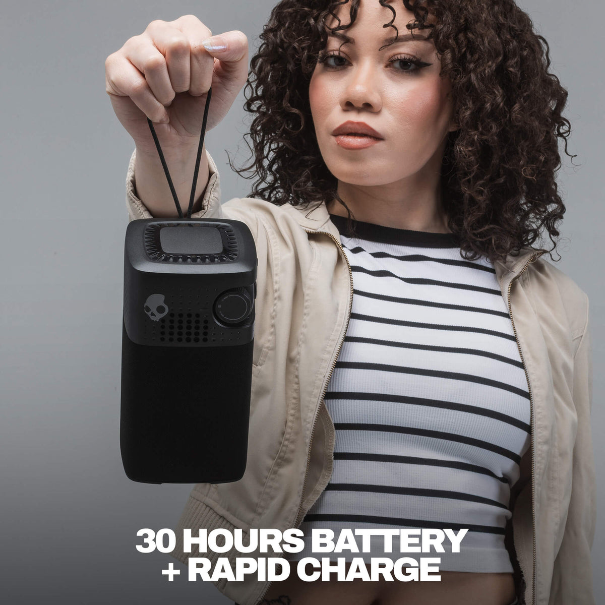 A woman holds up a black session 900 with a skull logo, wearing a beige jacket over a black and white striped shirt. Text at the bottom reads '30 HOURS BATTERY + RAPID CHARGE' against a gray studio background. | True Black