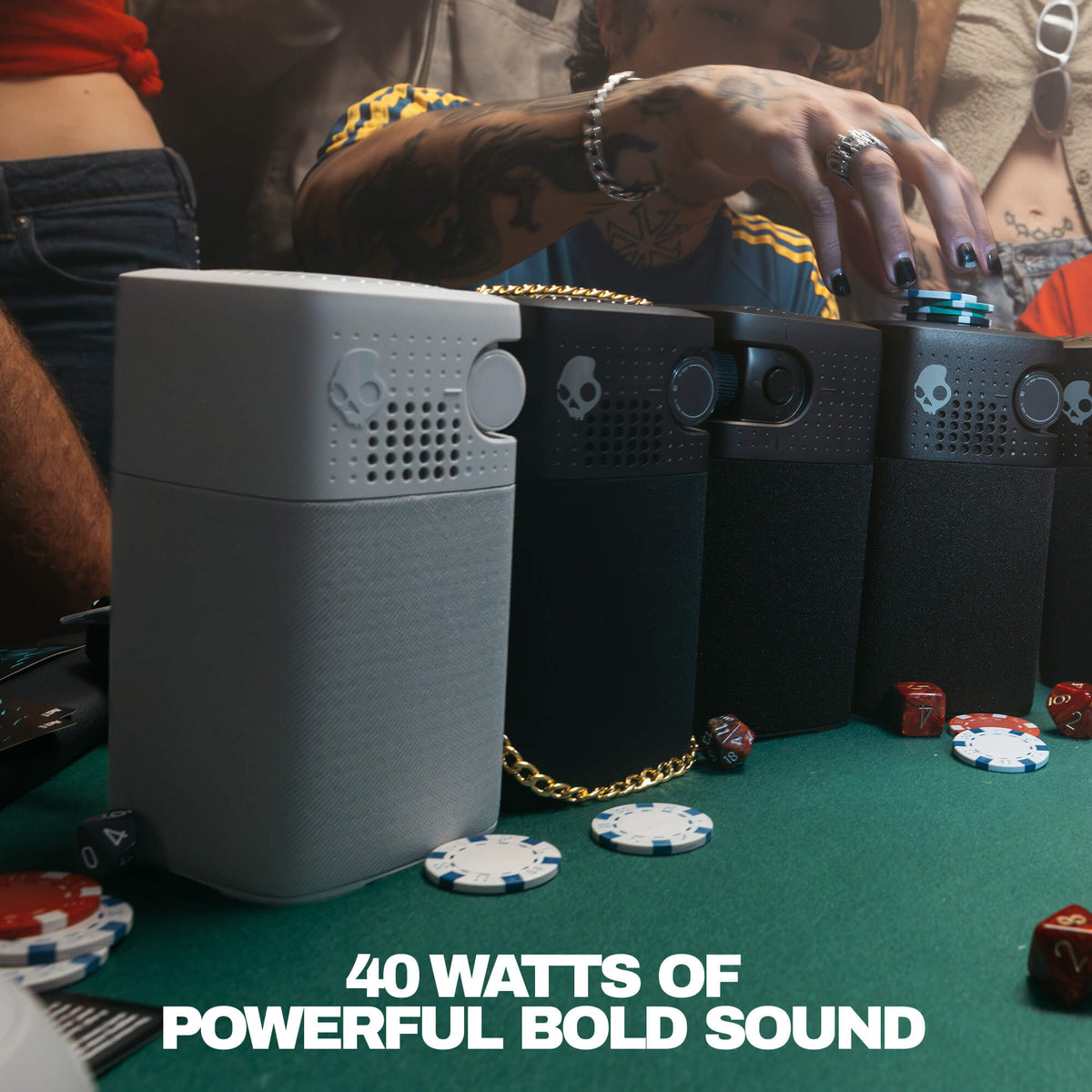 Three Skullcandy portable speakers in gray and black are lined up on a green felt table with poker chips and dice, with a person's tattooed hand reaching toward the devices. Text at the bottom reads '40 WATTS OF POWERFUL BOLD SOUND'. | Primer