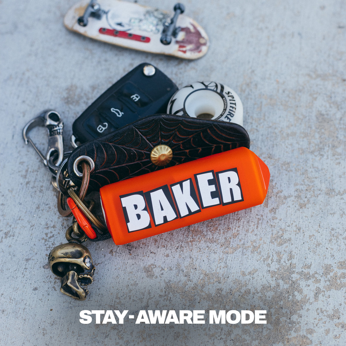 A close-up of a Skullcandy Dime 360 X Baker skateboards. The set includes a bright orange rectangular case with the word "BAKER" in bold white letters, a black spiderweb-patterned pouch, a black car key fob, a skateboard wheel, a skull charm, and metal hardware. | Skullcandy X Baker Skateboards