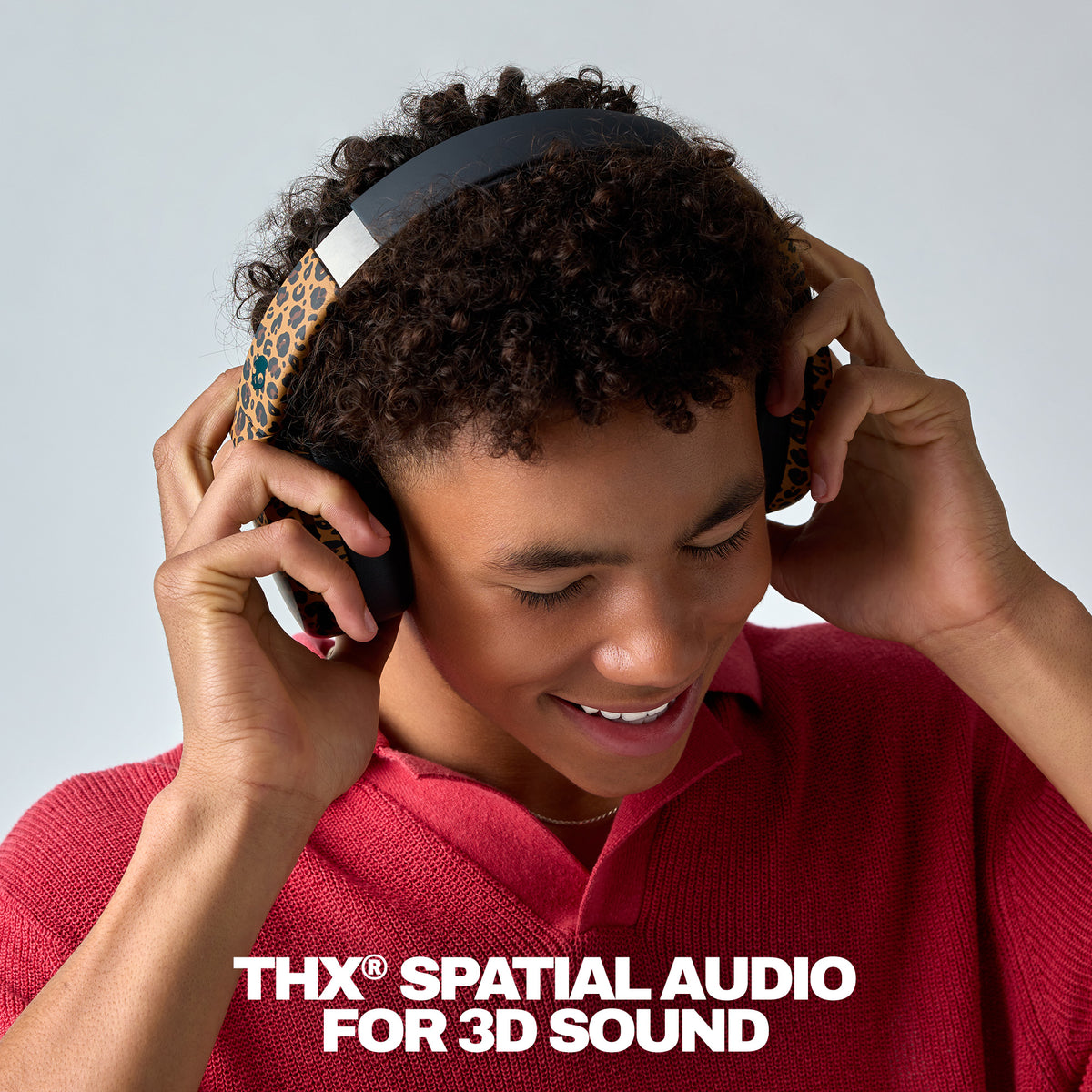 A young man with curly dark hair wearing over-ear headphones with a leopard-print pattern and a black headband, smiling while looking down. He is wearing a red knit polo shirt against a light gray studio background, with the text 'THX SPATIAL AUDIO FOR 3D SOUND' at the bottom.  | Leopard