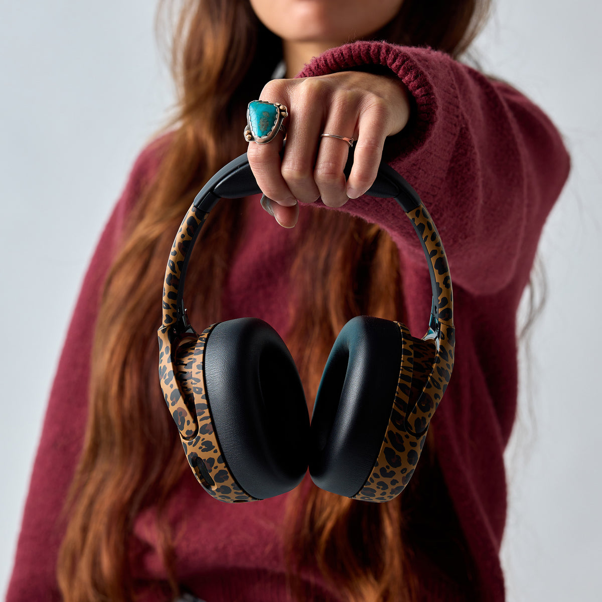 A person in a burgundy sweater holds a pair of over-ear headphones with black ear cushions and leopard-print fabric headband and ear cups against a light gray background. | Leopard