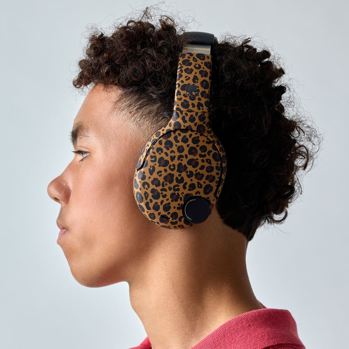 A side-profile view of a person with curly dark hair wearing over-ear headphones with a tan and black leopard print pattern, set against a plain light gray background. | Leopard