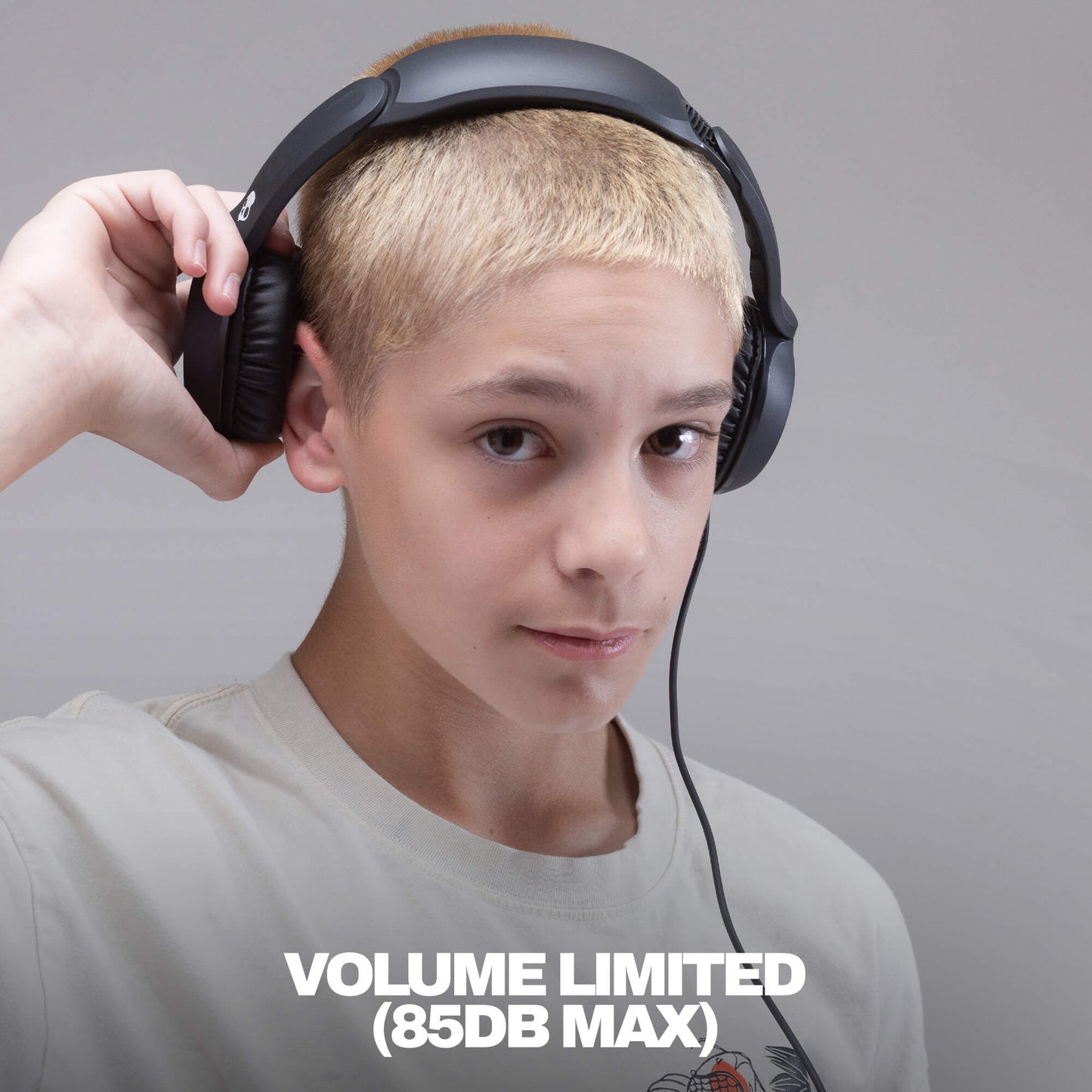 A person with short blond hair wearing black over-ear headphones and a light gray T-shirt, holding one earcup with one hand against a plain gray background. White text at the bottom reads 'VOLUME LIMITED (85DB MAX)'. | True Black