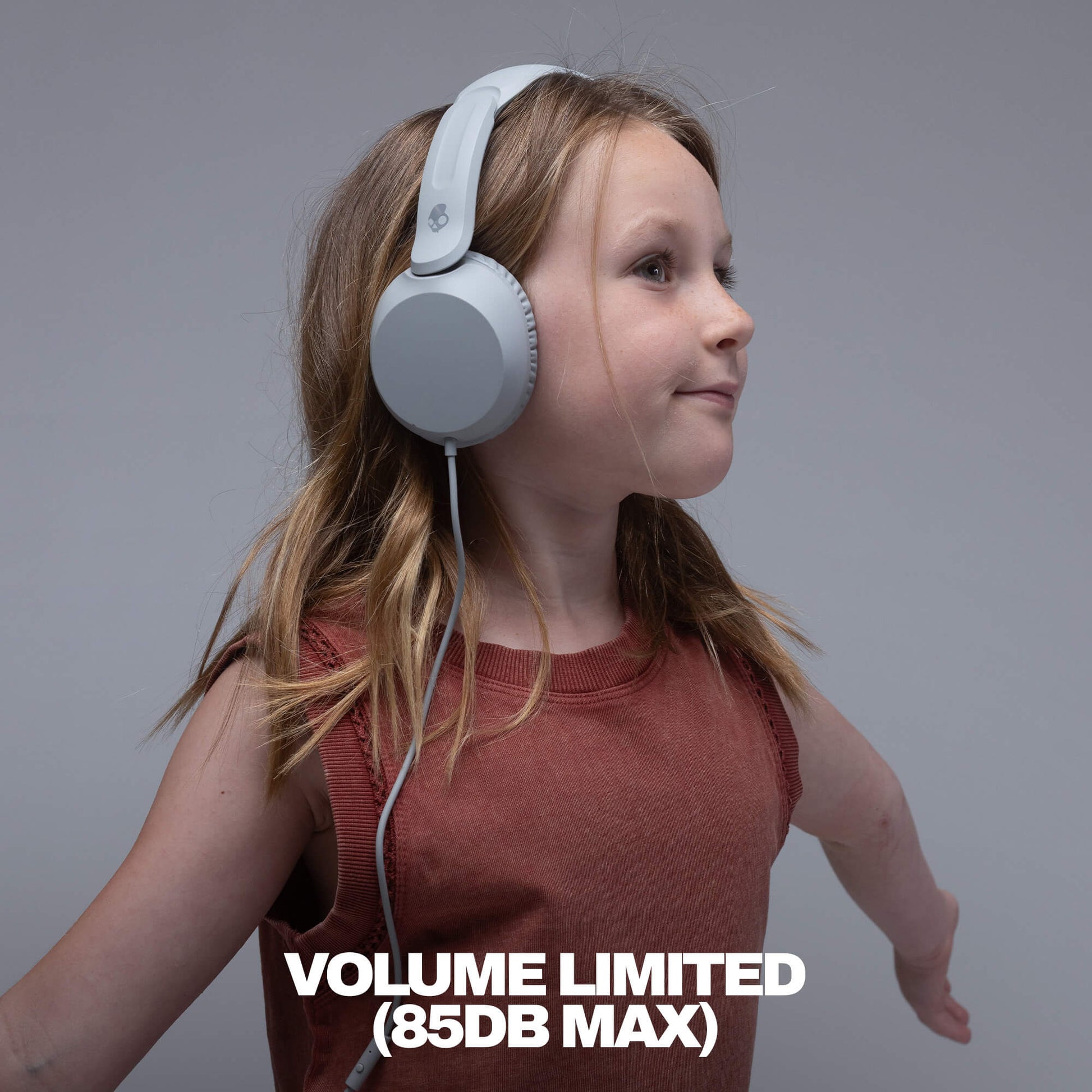 A young girl with blonde hair wearing light gray over-ear headphones and a rust-colored sleeveless top, shown in profile against a solid gray background with the text 'VOLUME LIMITED (85DB MAX)' at the bottom.  | Cement