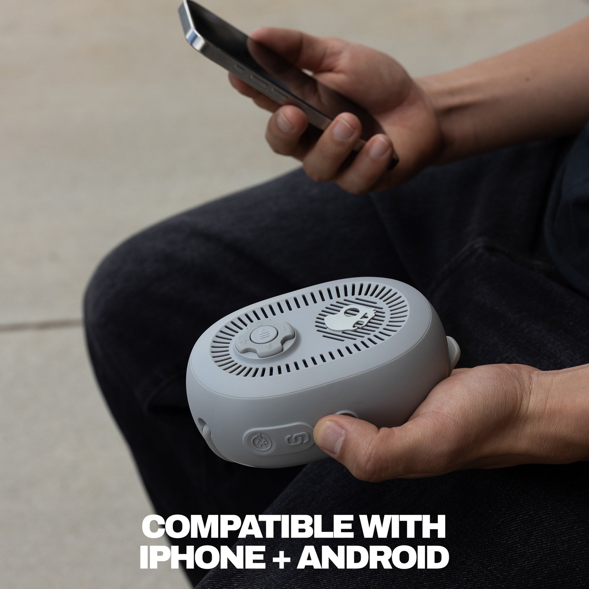 Person holding a small grey portable speaker with a phone, sitting on a light-colored floor. | Cement