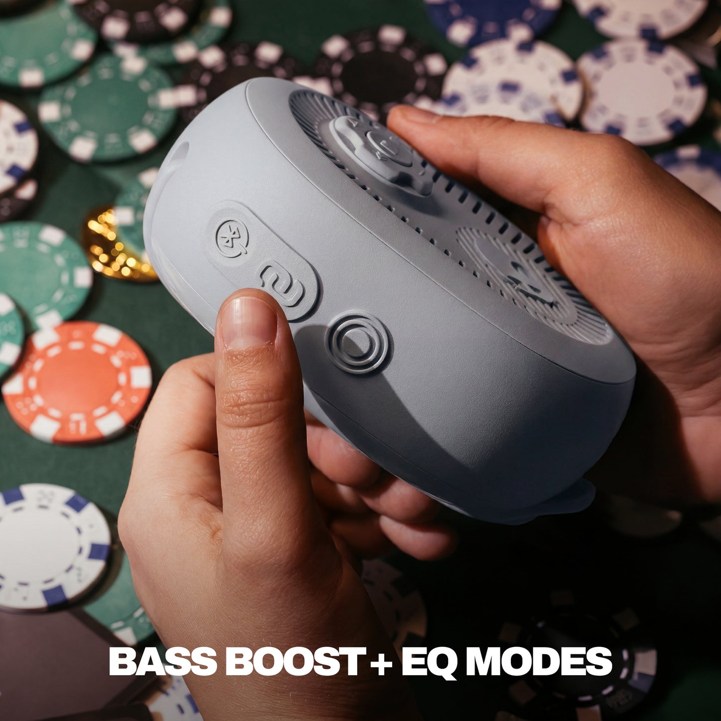 Person holding a gray portable speaker with buttons, surrounded by poker chips on a green felt surface. | Cement