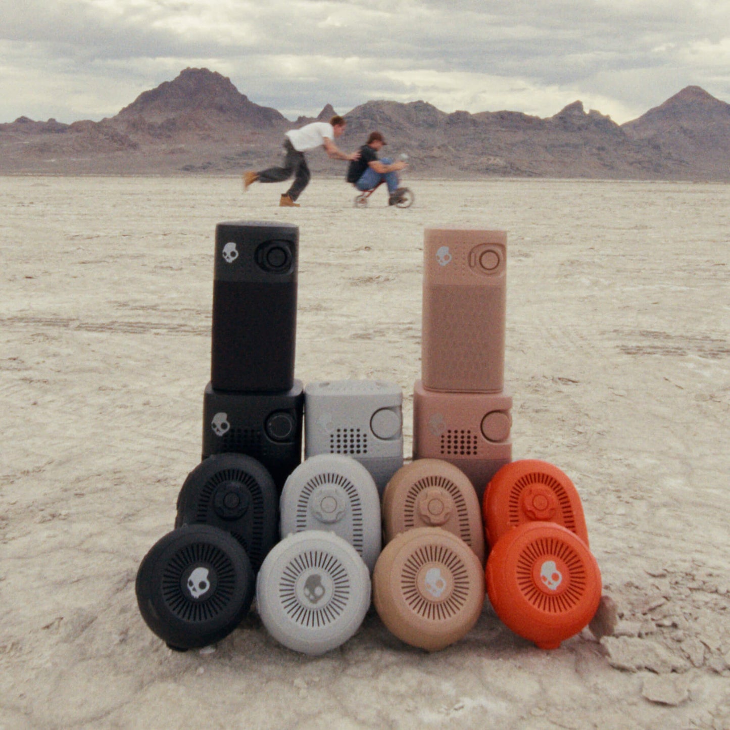 A set of portable speakers in multiple colors (black, gray, beige, orange) arranged in a pyramid on a desert flatland with mountains in the background. Two people are blurred in the background riding a bicycle. | Cement